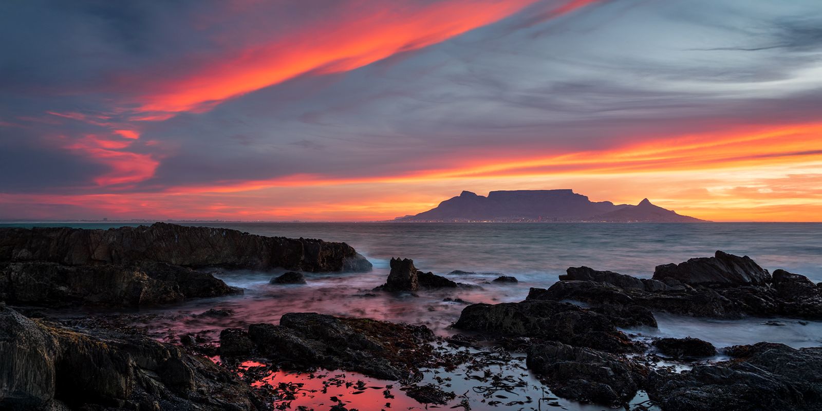 Cape Town, South Africa sunset view of Table Mountain with rocky shoreline and vibrant sky.