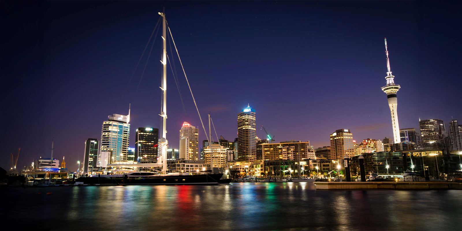 Auckland, New Zealand harbor skyline at night with illuminated buildings and sailboats.