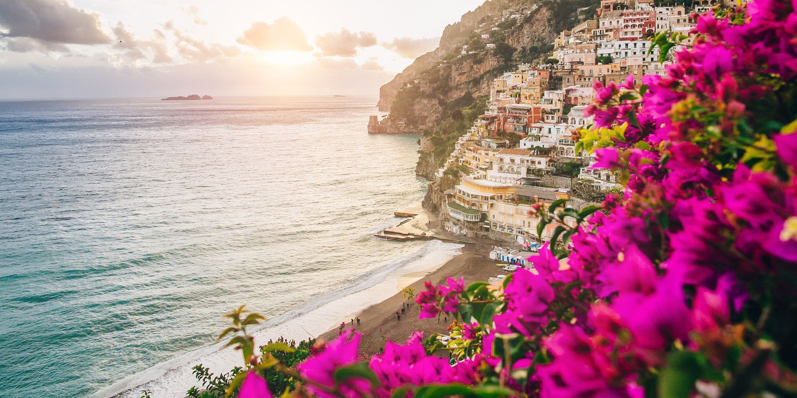 Colorful cliffside buildings of Positano on Italy’s Amalfi Coast at sunset, framed by vibrant pink bougainvillea overlooking the sea.!''