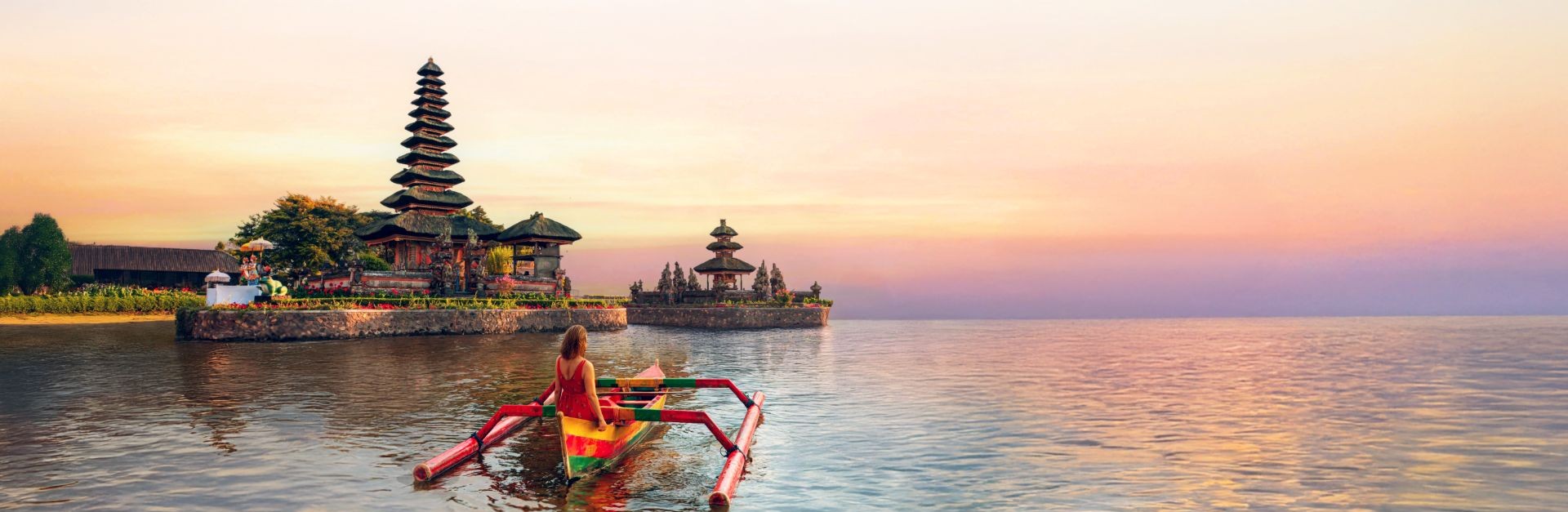 Woman in a traditional wooden boat at sunset facing Pura Ulun Danu Beratan temple on a lake in Bali.