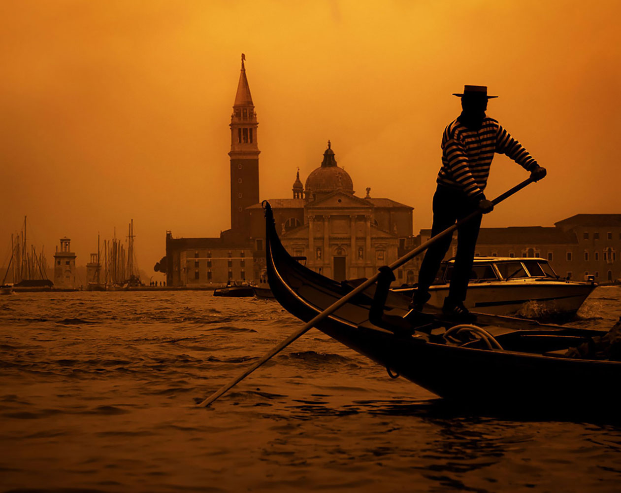 A gondolier guides his gondola through a Venice canal in the orange glow of sunset.