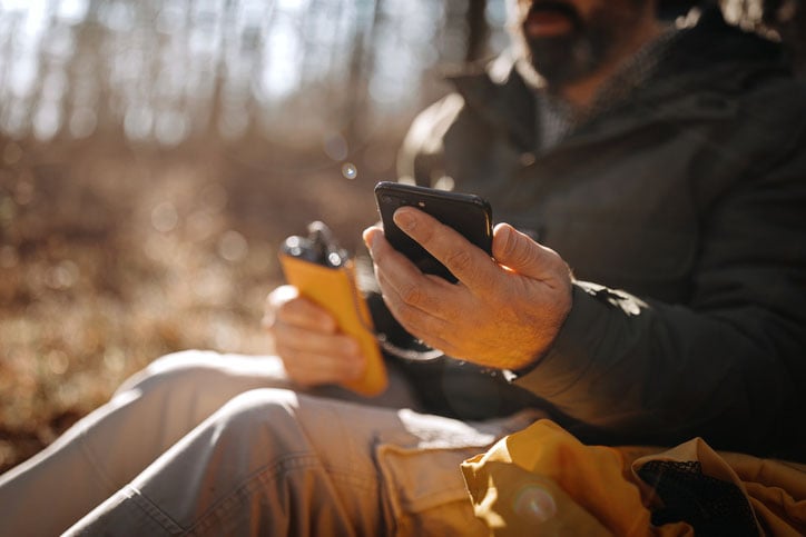 Man sitting in woods and charging mobile phone with power bank during beautiful day