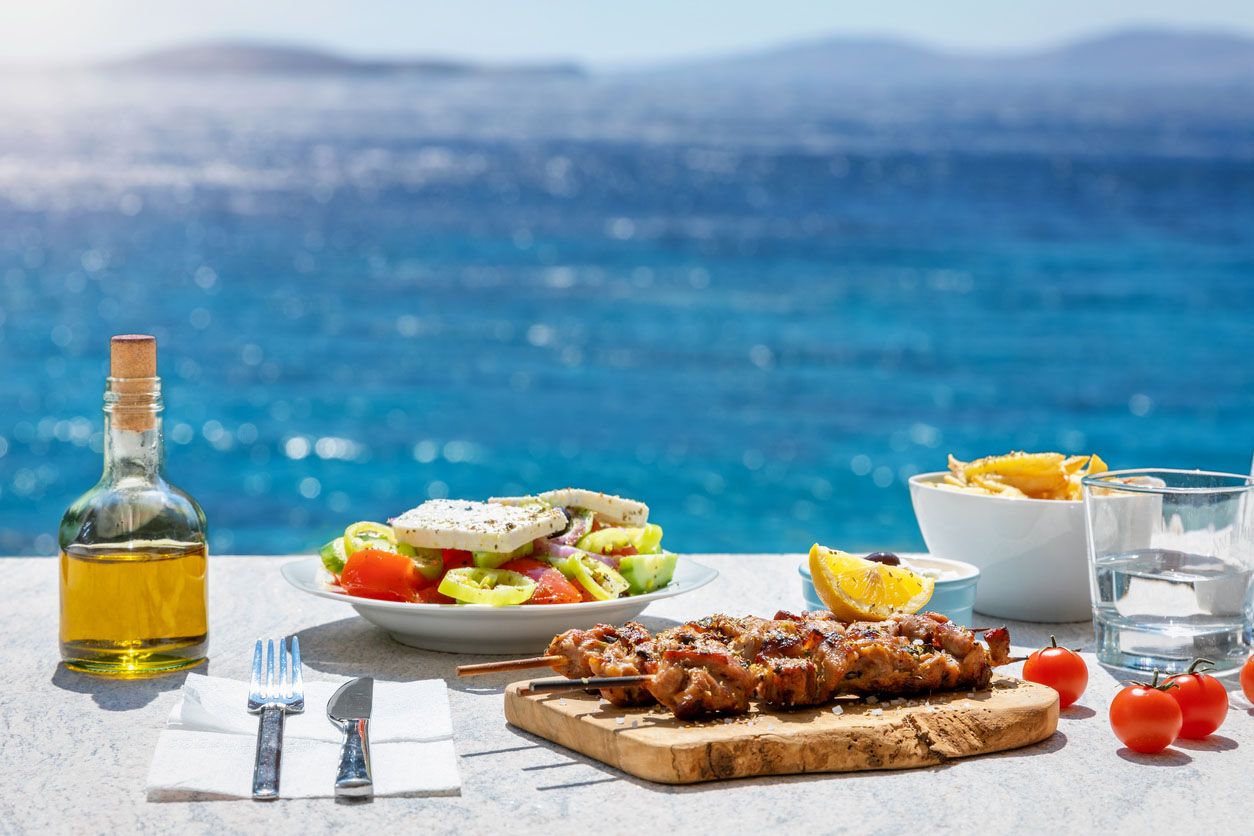 Farmers salad and souvlaki skewers beside a bottle of Greek olive oil in front of the Aegean sea