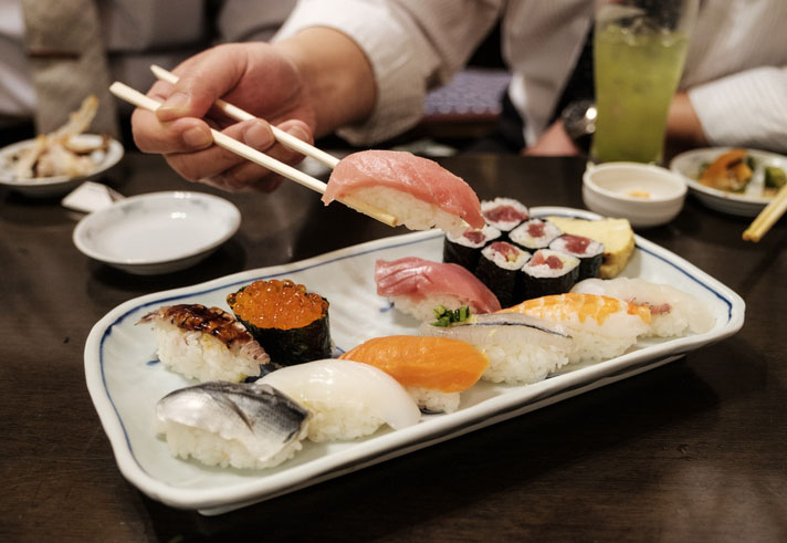 Chopsticks holding a piece of Tuna sushi above a full plate of various nigiri and maki sushi.