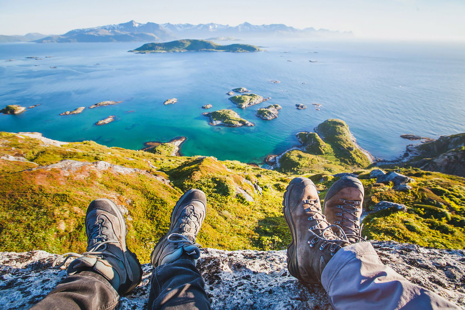 Two hikers overlook a lake in the Norwegian Fjords from a mountain top, with their hiking shoes in the foreground.