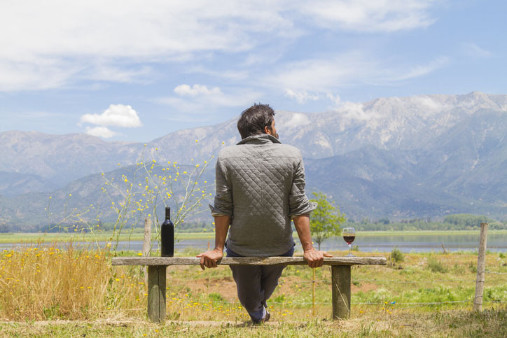 A man sits on a bench with a bottle of wine beside him and the mountains in the background