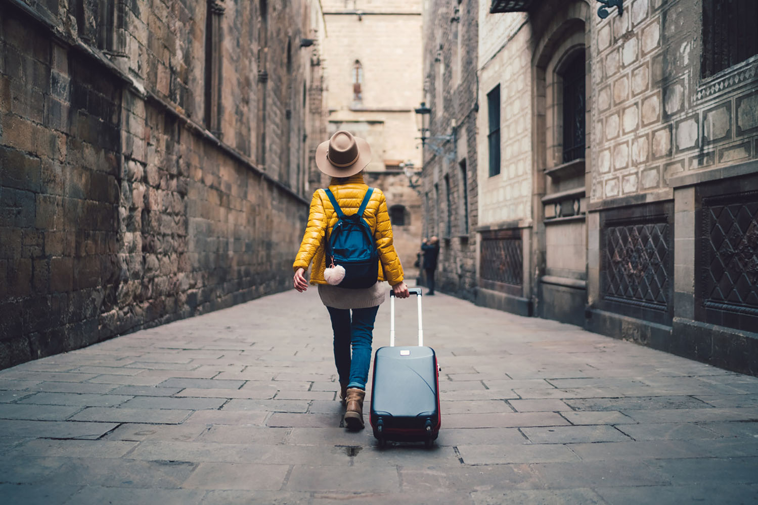 A woman in yellow walks down a cobblestone street with luggage