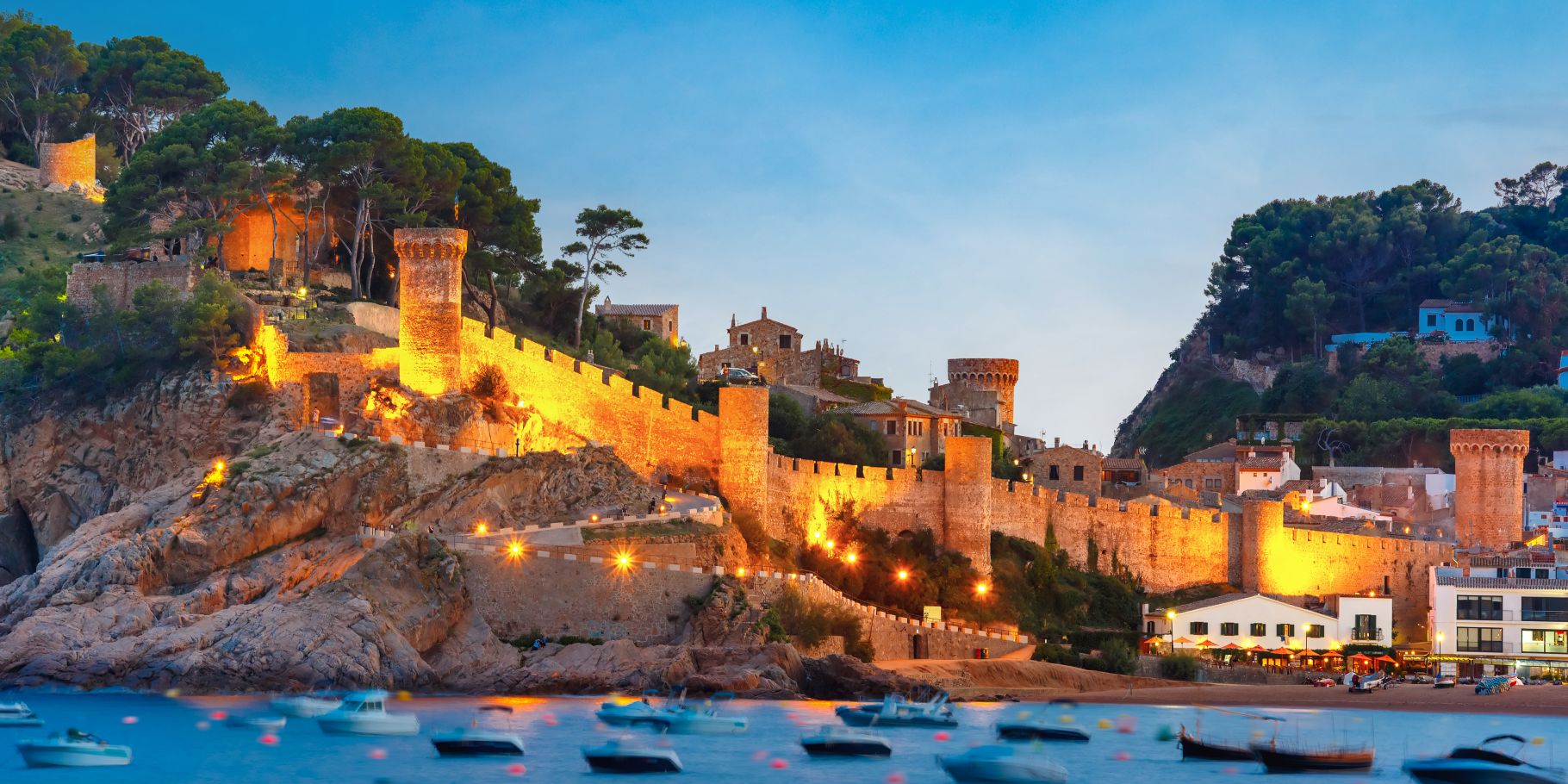 Medieval castle of Tossa de Mar in Costa Brava, Spain illuminated at dusk with boats in the bay.