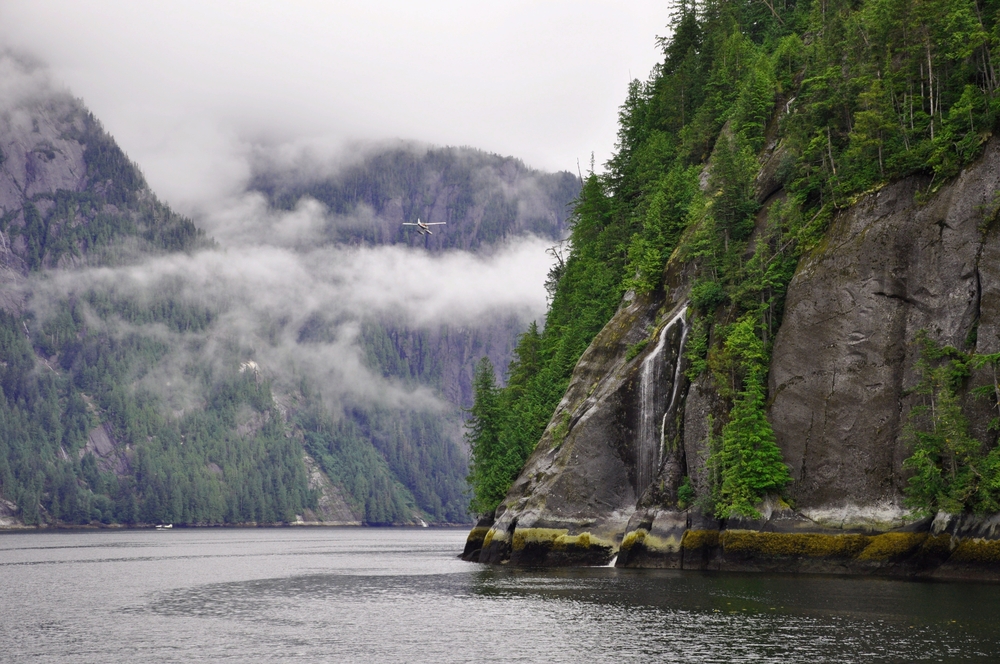 Misty Fjords National Monument with sheer granite cliffs, cascading waterfalls, and a floatplane flying through low coastal mist in Alaska.